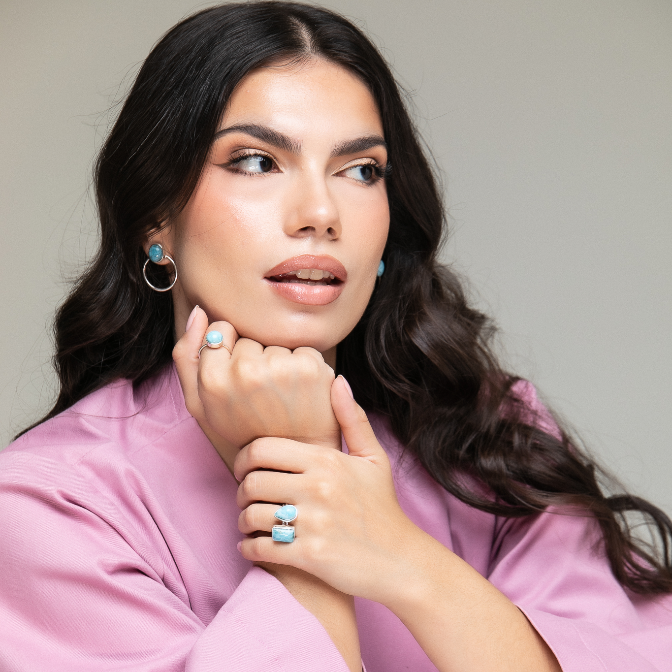 Woman wearing a pink top, styled with silver Larimar hoop earrings and multiple bold Larimar rings, gazing off-camera with soft waves and natural makeup.