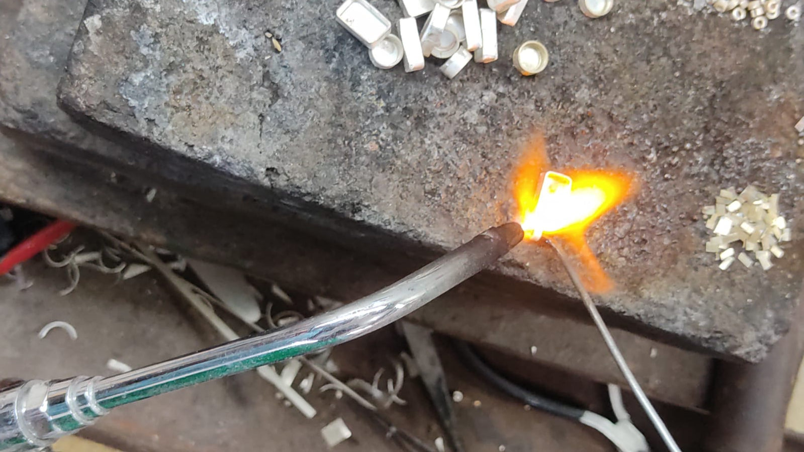 Silversmith using a torch to solder a handcrafted jewelry piece on a workbench, surrounded by small silver components.