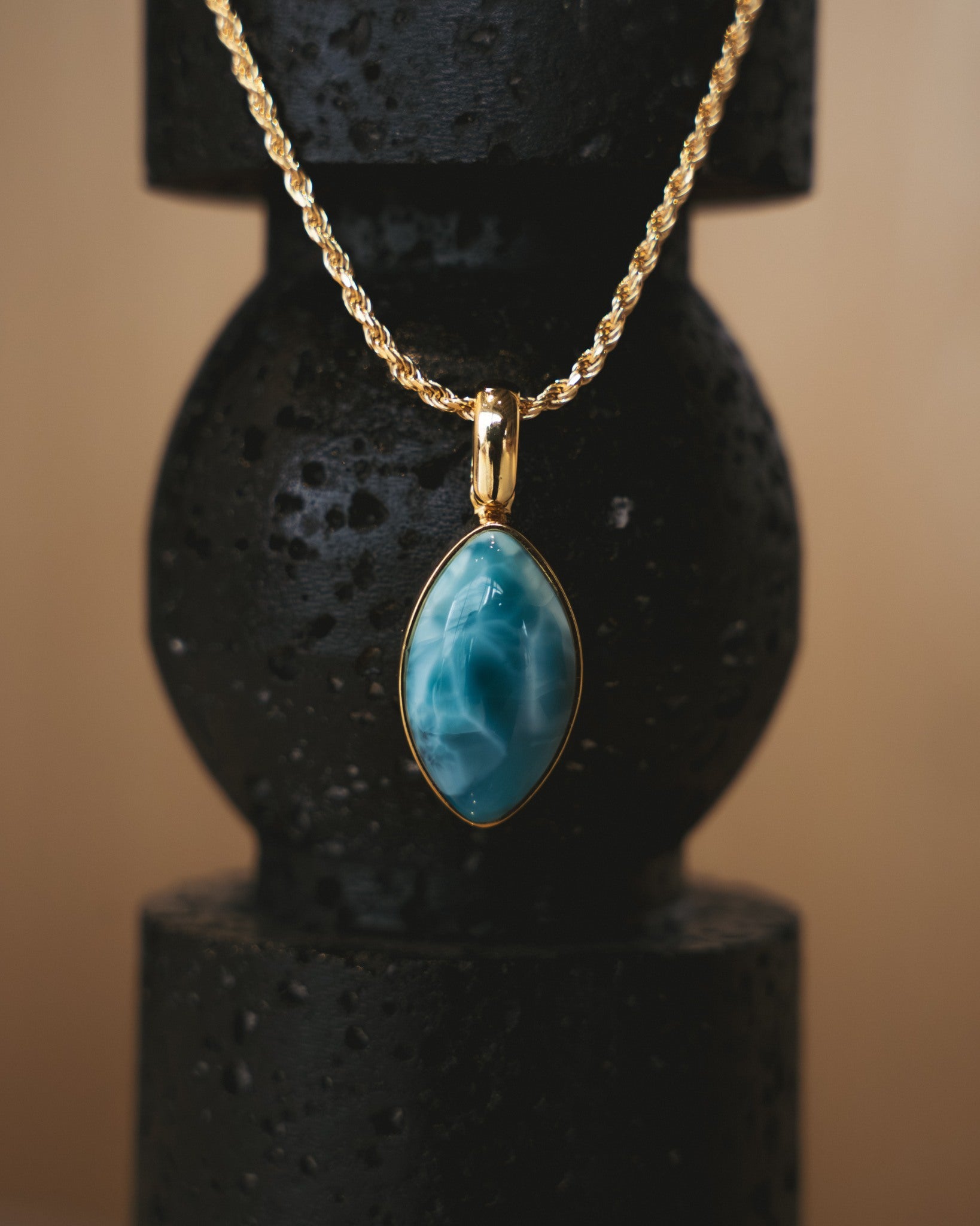 Gold necklace with a blue Larimar pendant on a black stand against a beige background.