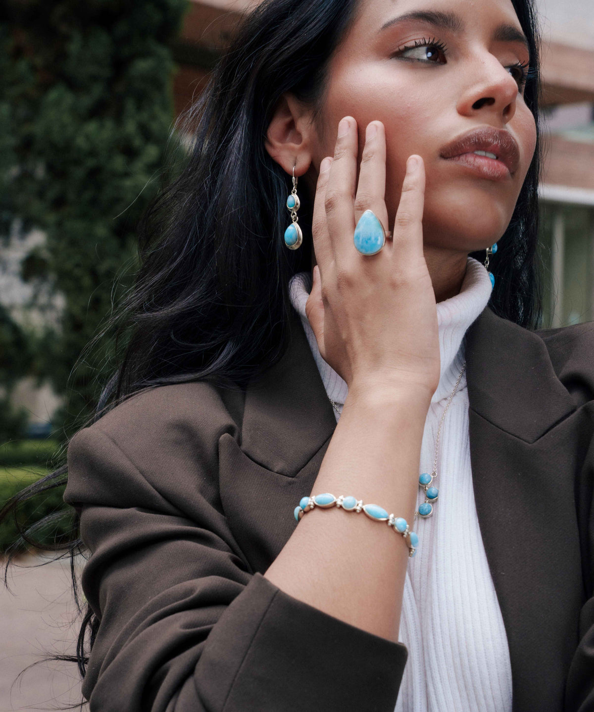 A woman wearing a matching Larimar and silver jewelry set, including a ring, earrings, and bracelet, styled with a brown blazer and white turtleneck.
