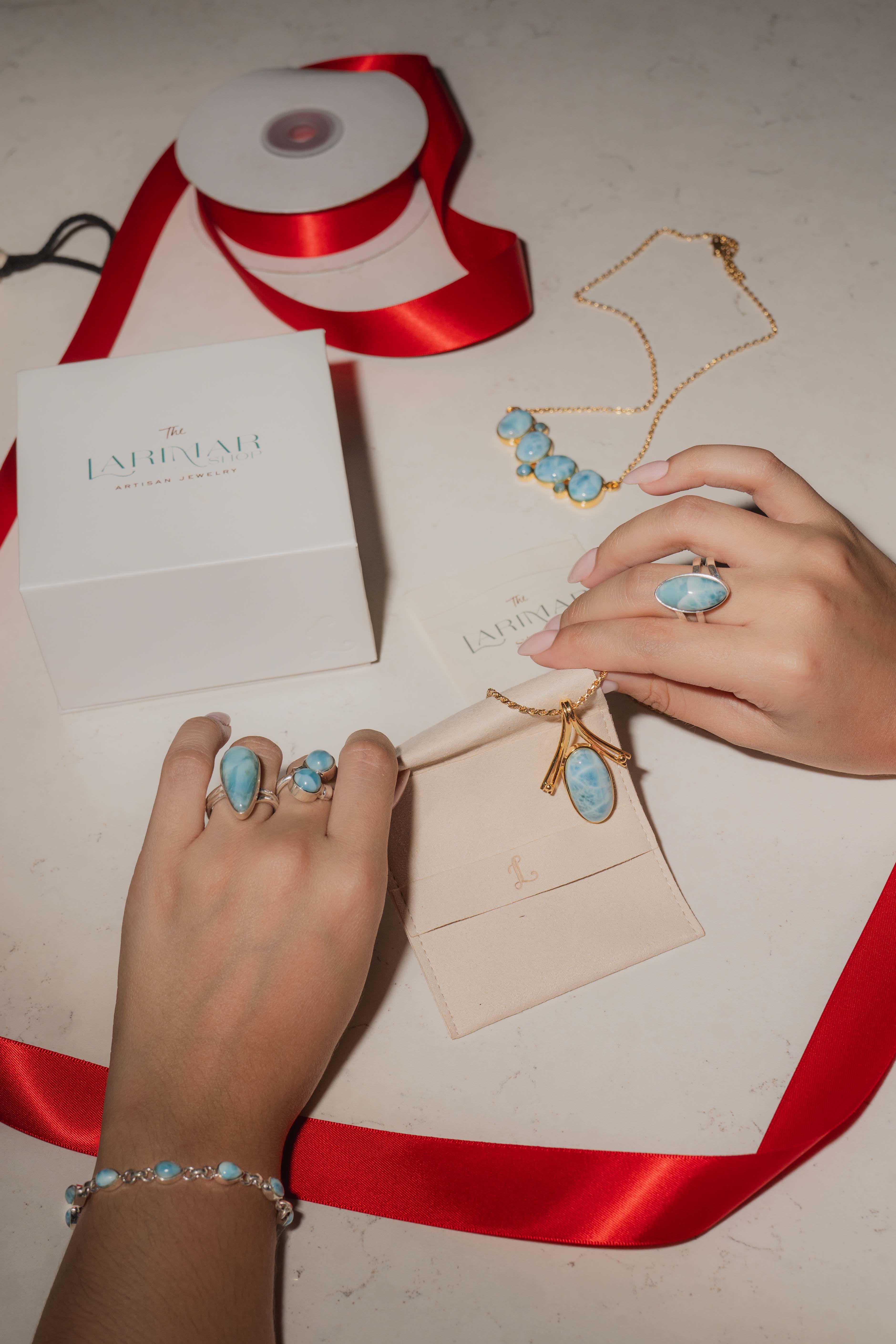 Hands arranging beautifully wrapped holiday gifts and showcasing one-of-a-kind Larimar jewelry pieces, including rings, bracelets, necklaces, and earrings, accented with festive red ribbons and gift boxes.