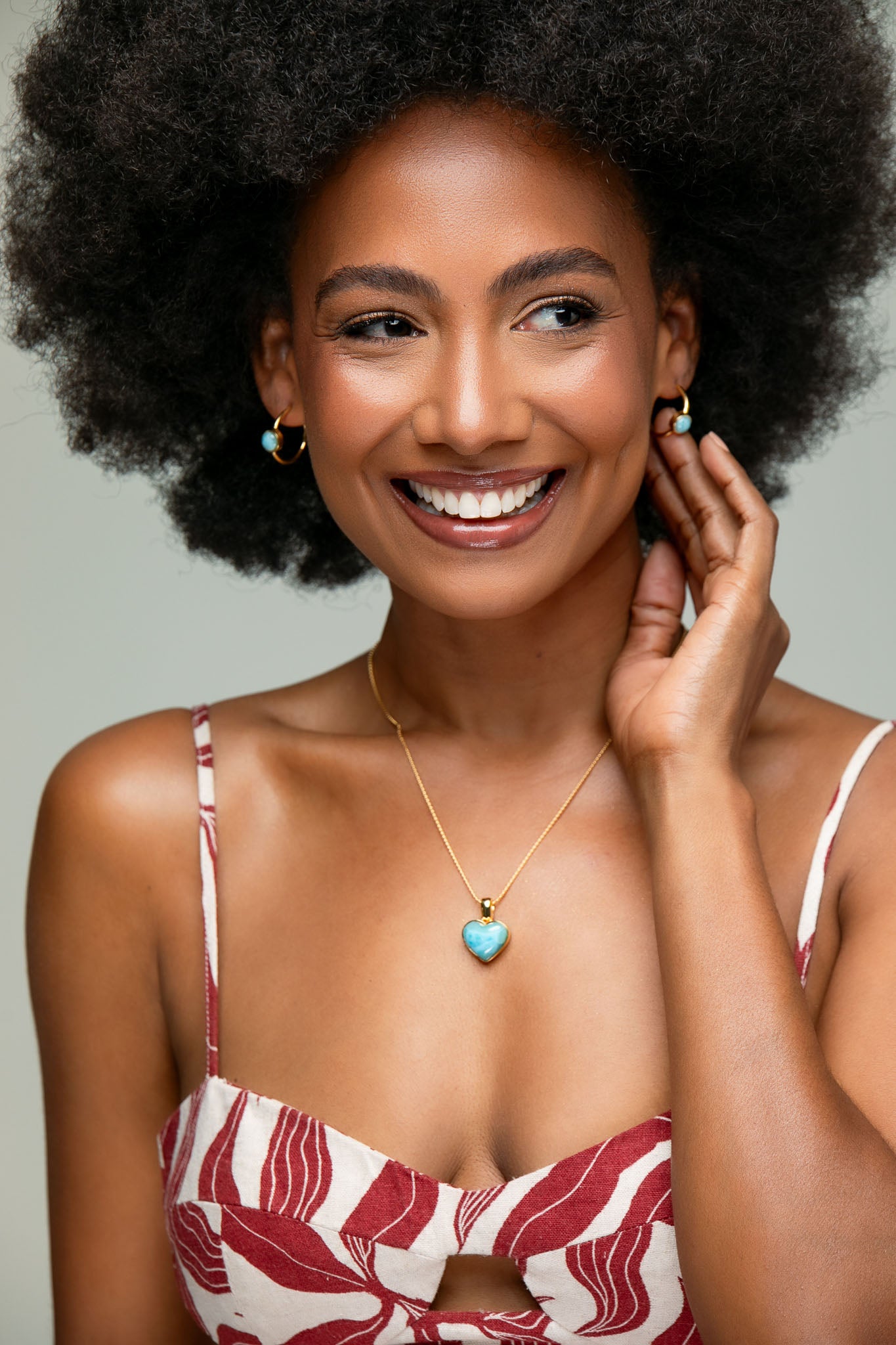 Woman with an afro wearing a red and white patterned top, Larimar gold necklace, and earrings against a gray background