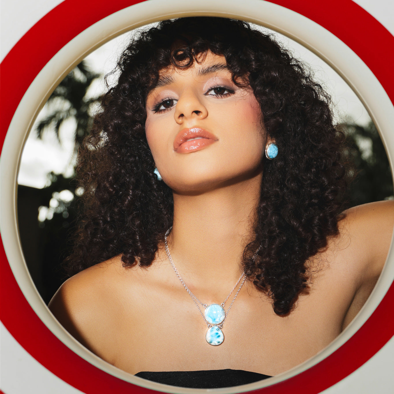 Woman with curly hair wearing a Larimar necklace with a blue pendant, framed by a red and white circular border.