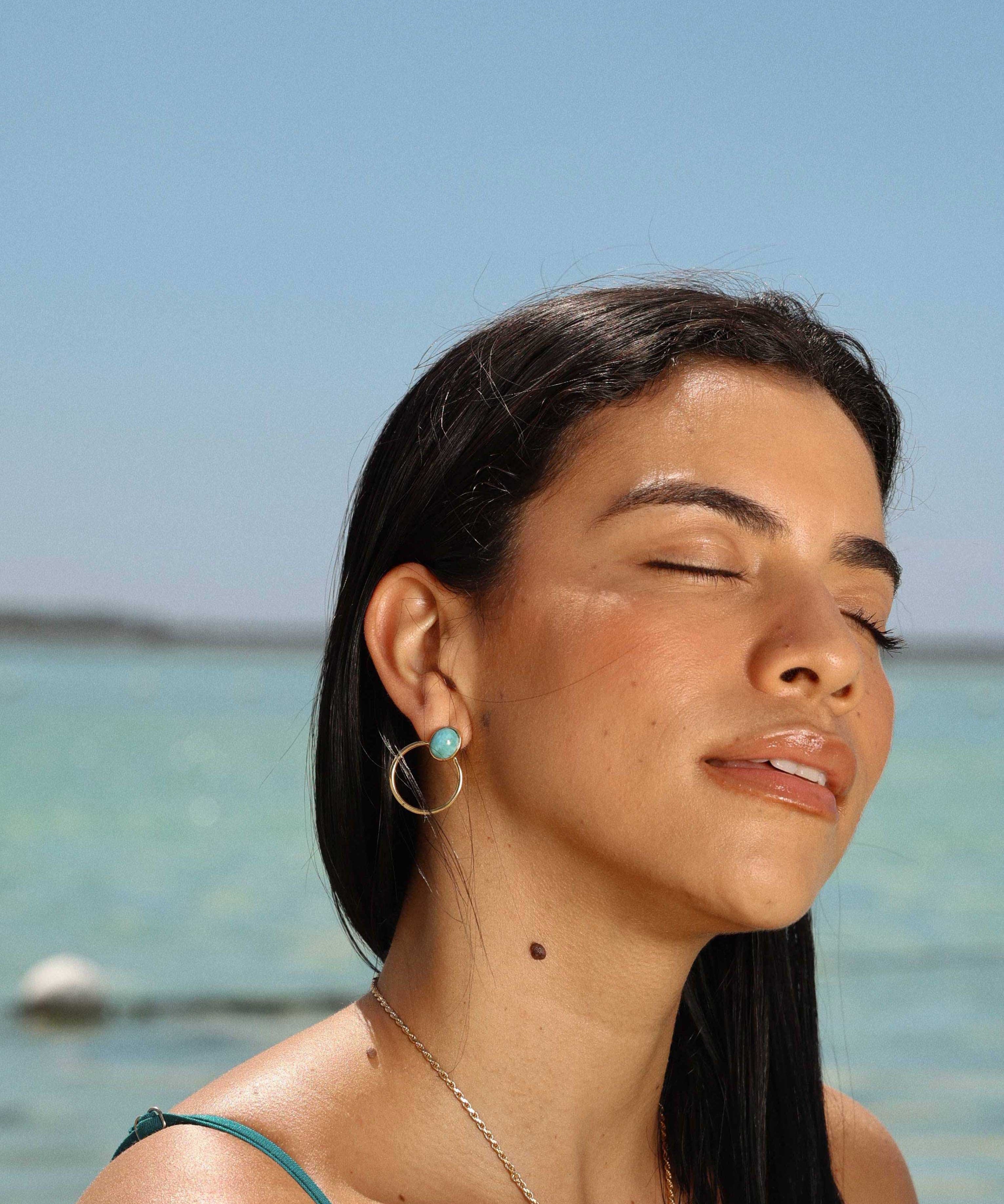 Woman with closed eyes standing by a body of water under a clear blue sky wearing The Larimar Shop jewelry 