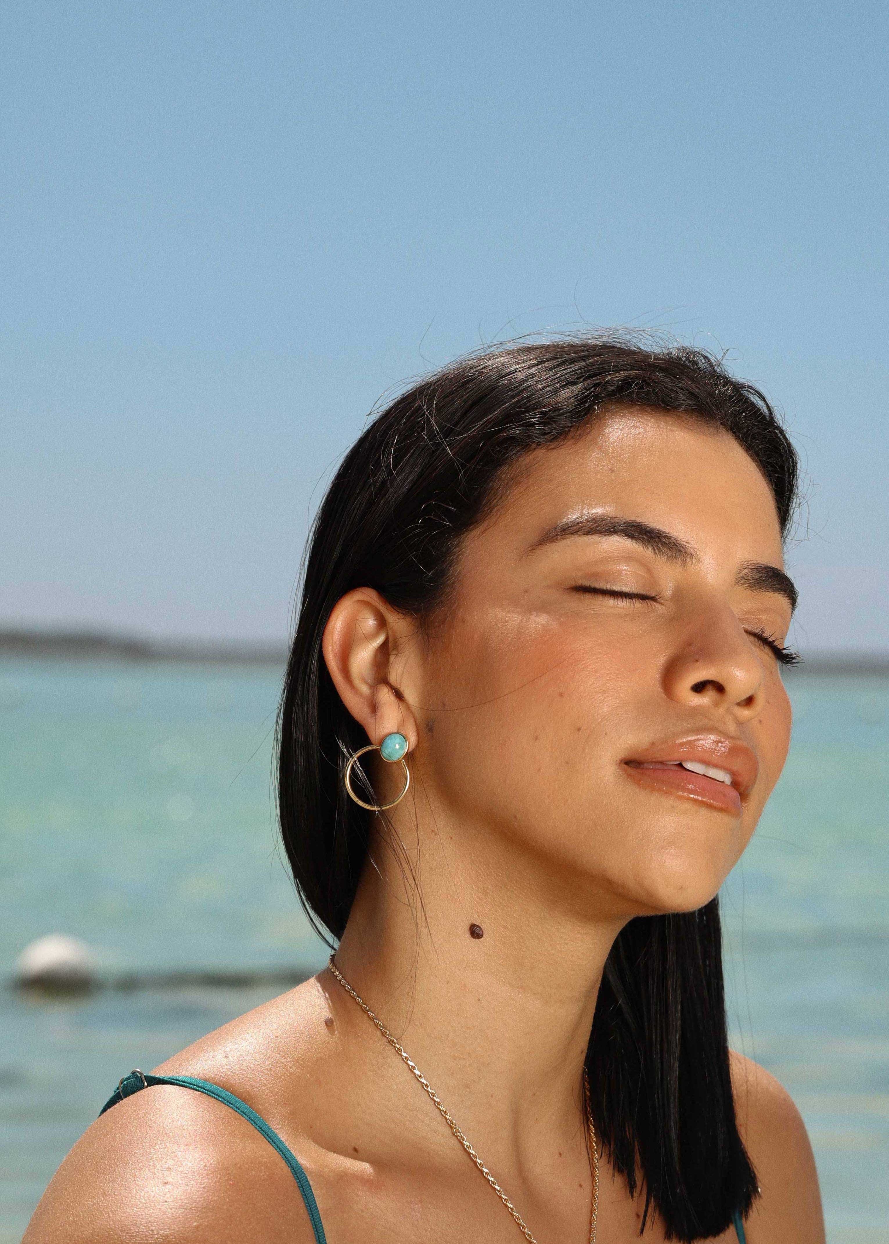 Woman with closed eyes standing by a body of water under a clear blue sky wearing The Larimar Shop jewelry 