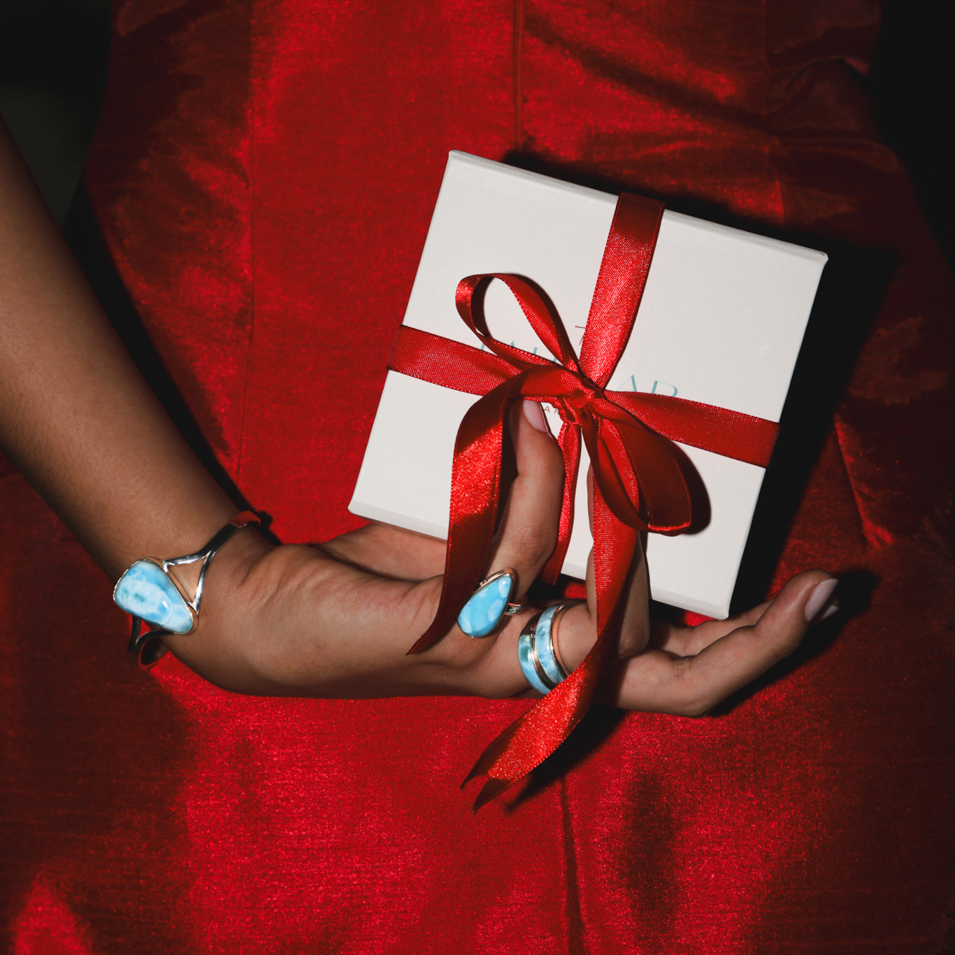 Person wearing Larimar gold jewelry holding a gift box with a red ribbon on a red background