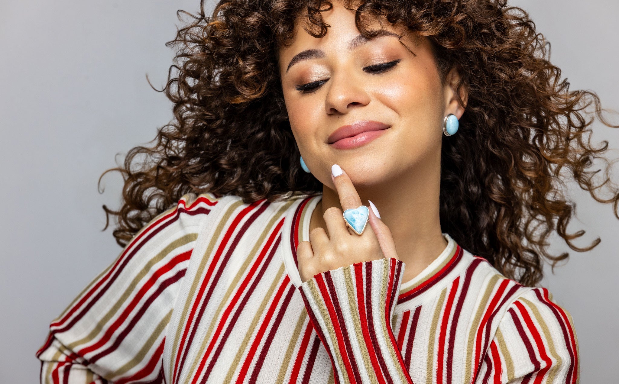 Woman with curly hair wearing a Larimar ring and earrings with a striped shirt against a gray background.