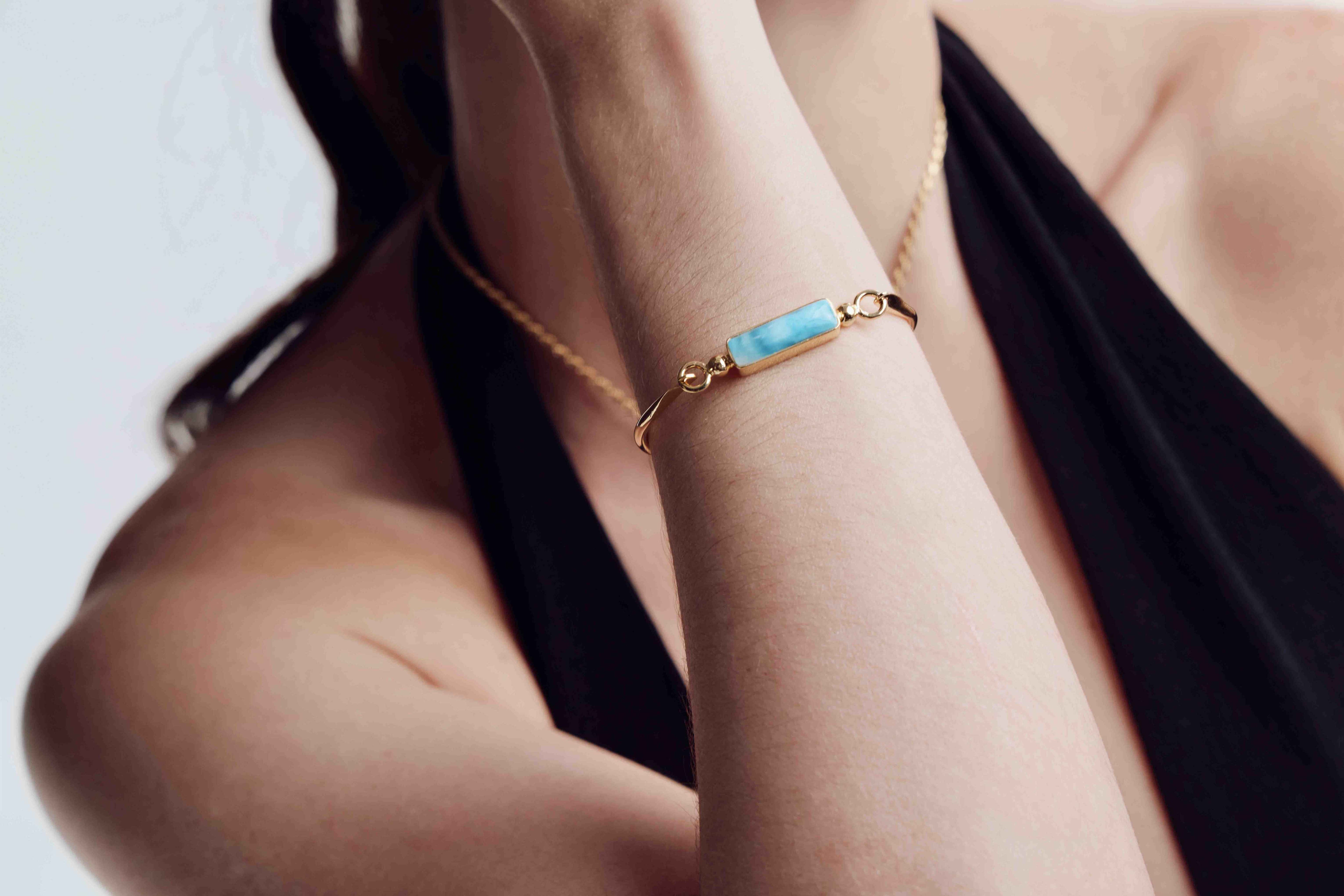 Close-up of a person wearing a gold bracelet with a blue Larimar stone on a neutral background
