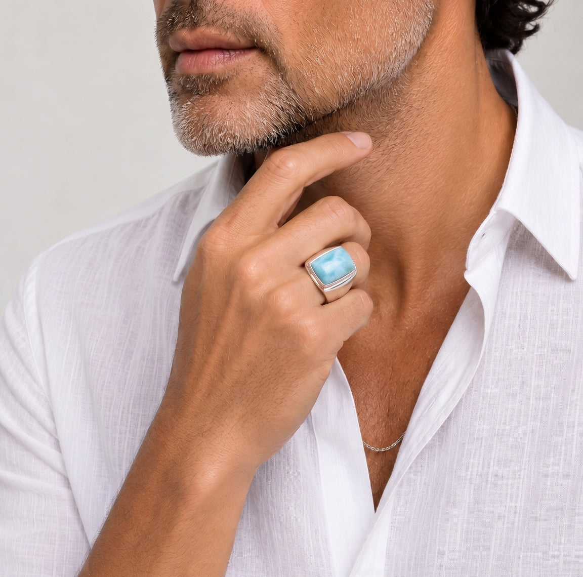 Man wearing a white shirt with a blue Larimar ring on his finger, touching his chin.