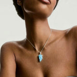 Close-up of a person wearing a silver chain with a blue wing Larimar pendant against a neutral background