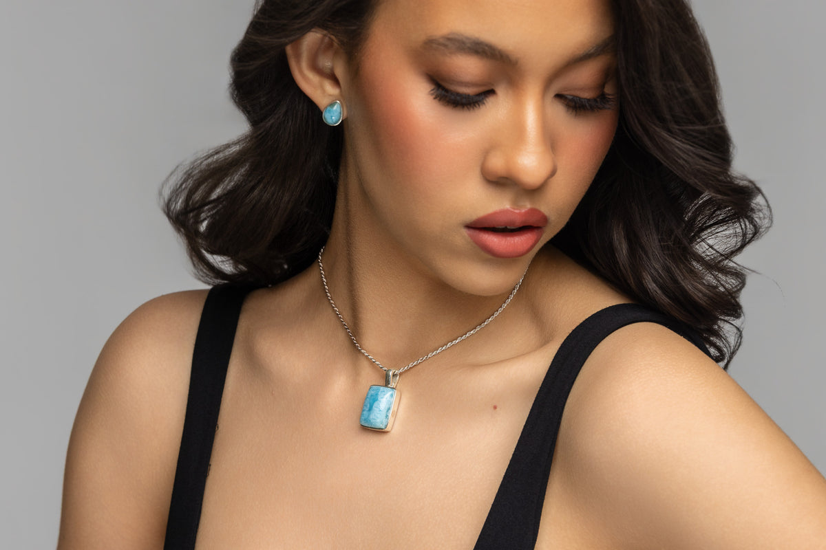 A woman wearing a silver-toned necklace with a rectangular Larimar-blue stone pendant and matching stud earrings, shown against a plain gray studio background.