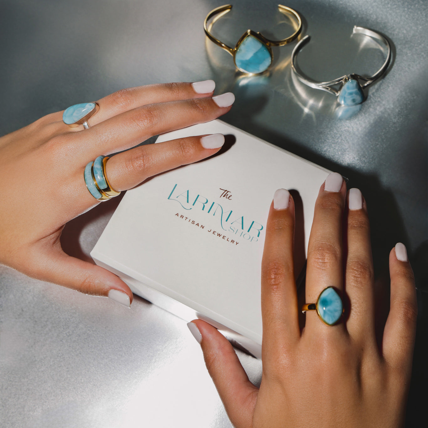 Hands wearing Larimar gold and silver rings with 'The Larimar Shop' jewelry box in the background.