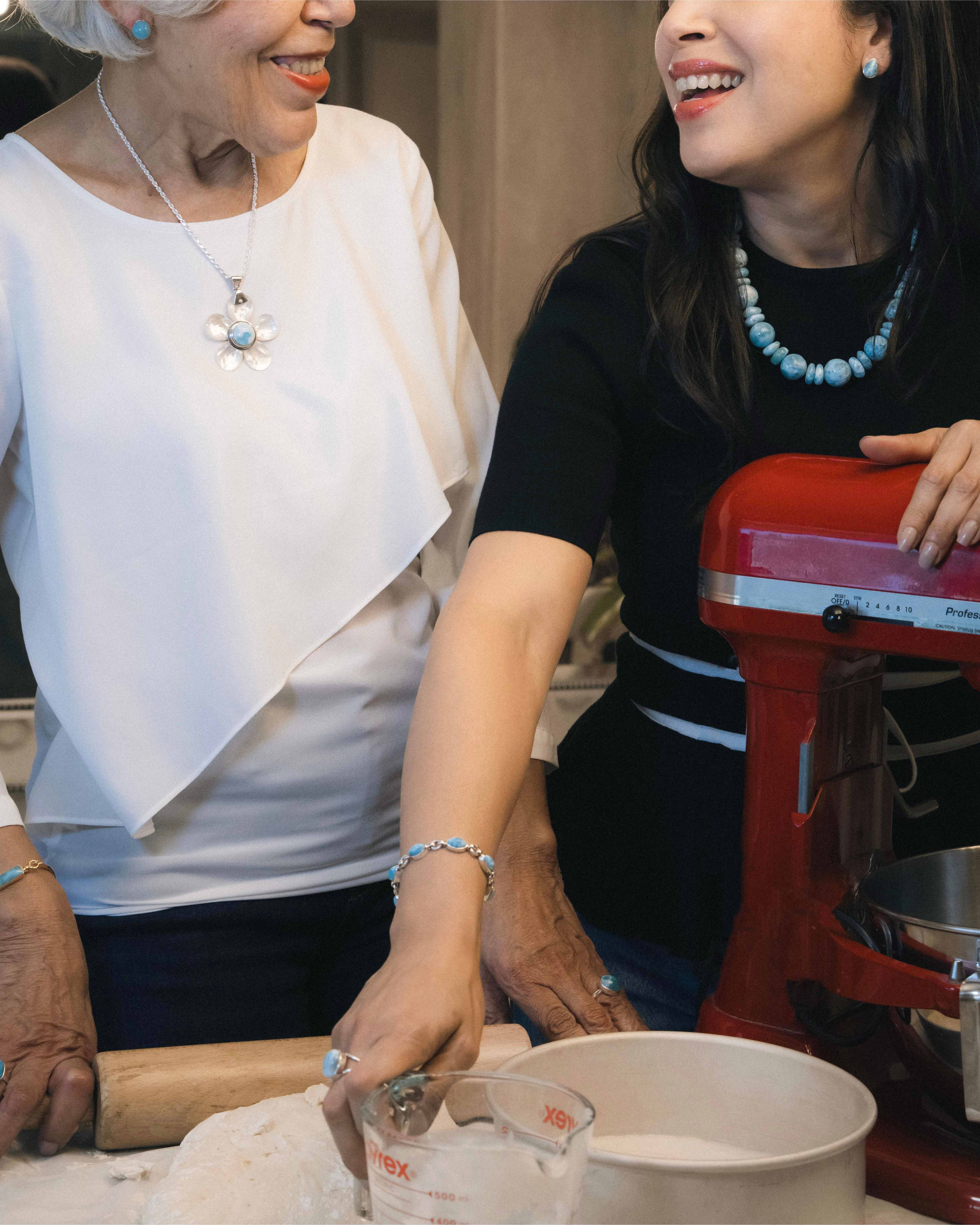 Two women wearing Larimar jewelry from The Larimar Shop baking together, one using a red stand mixer and the other with a rolling pin.