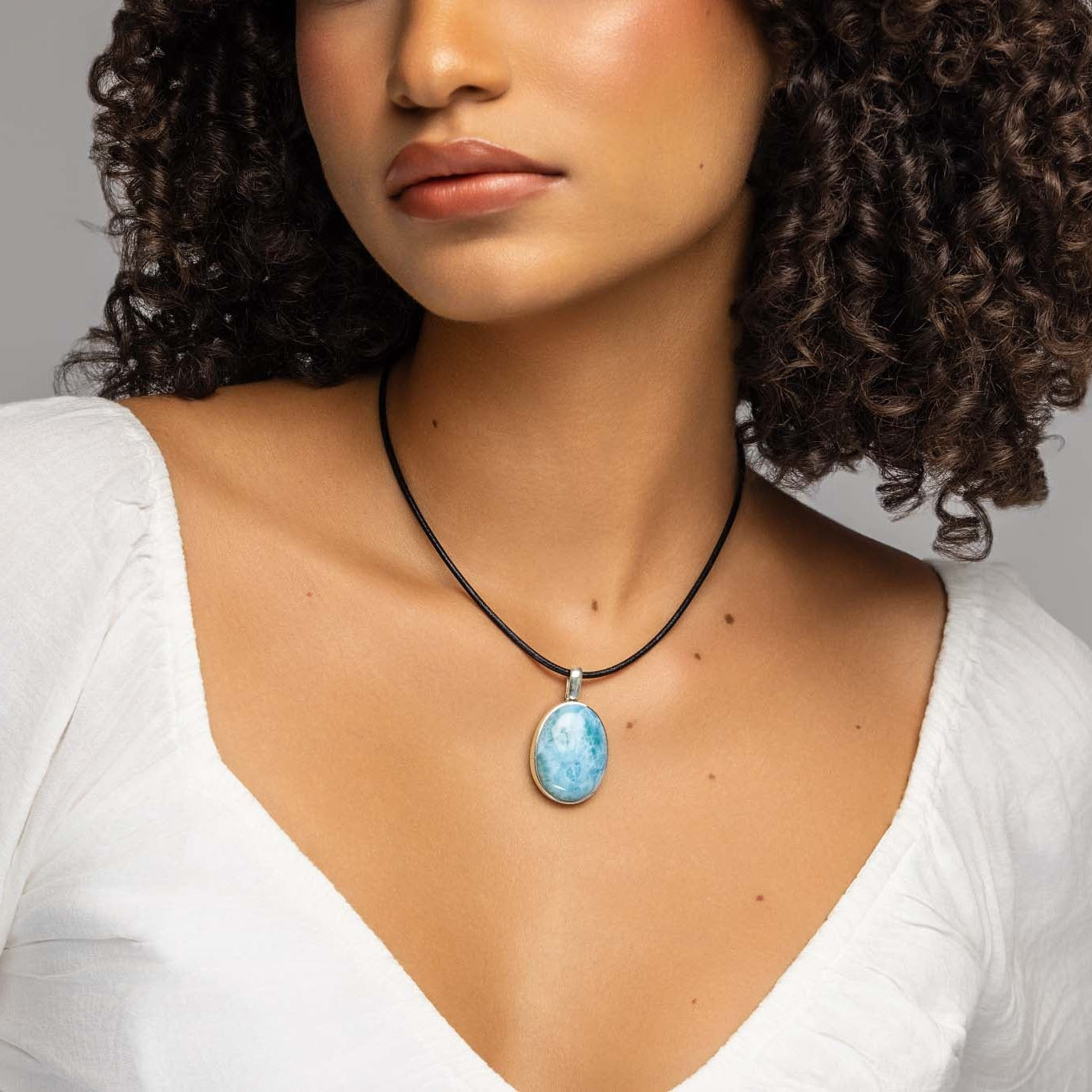A close-up of a woman wearing a black cord necklace with an oval light blue Larimar stone pendant, paired with a white top against a neutral gray background.